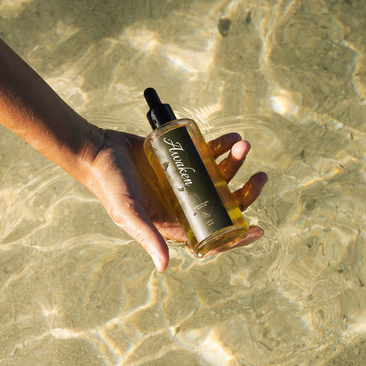 A hand holds a bottle of hair oil over clear, sunlit shallow water. The bottle is transparent with golden-amber liquid and a black dropper top, with a dark label featuring white script text.