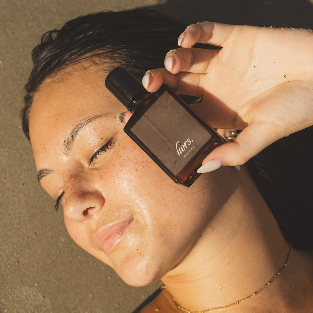 A woman lying on sand holds a small dark brown bottle near her face. The bottle has a black cap and a white script logo that appears to read 'leo'.