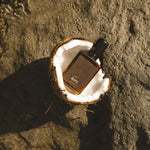 A small amber glass bottle with a black cap rests inside a halved coconut shell on a dark textured rock surface, with shallow water and sunlight visible in the upper area.