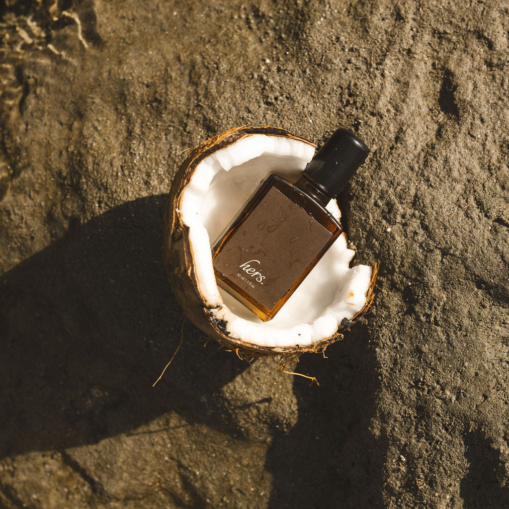 A small amber glass bottle with a black cap rests inside a halved coconut shell on a dark textured rock surface, with shallow water and sunlight visible in the upper area.