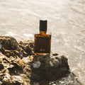 A small amber glass perfume bottle with a black cap and a dark label reading 'hors' is placed on a wet rock at the edge of shallow water.