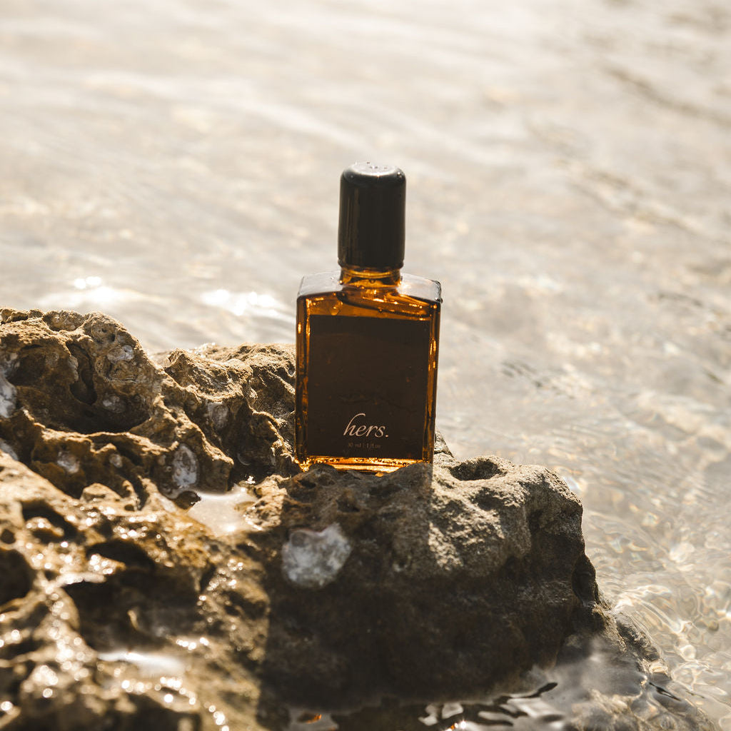A small amber glass perfume bottle with a black cap and a dark label reading 'hors' is placed on a wet rock at the edge of shallow water.